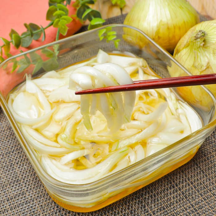 ガラス容器に入った玉ねぎのスライスを箸で持ち上げた様子。玉ねぎが背景に置かれている。