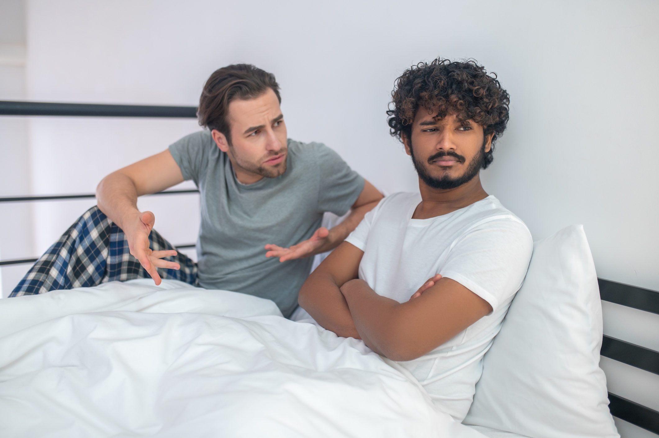 Two people in a discussion, one gesturing with hands, sitting on a bed with white sheets