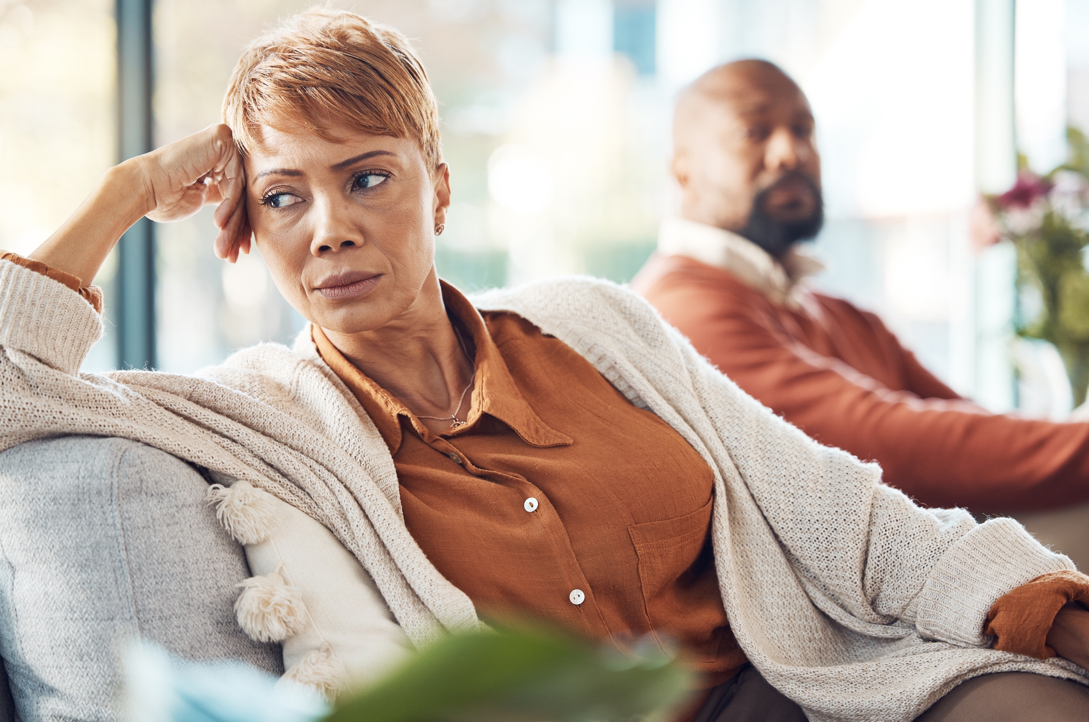 Woman in a cozy sweater looks contemplative while sitting on a sofa; a man sits in the background, also appearing thoughtful