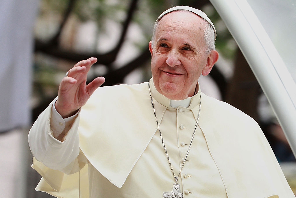 A smiling person in religious attire waves, wearing a robe, a white cap, and a cross necklace. Trees are visible in the background