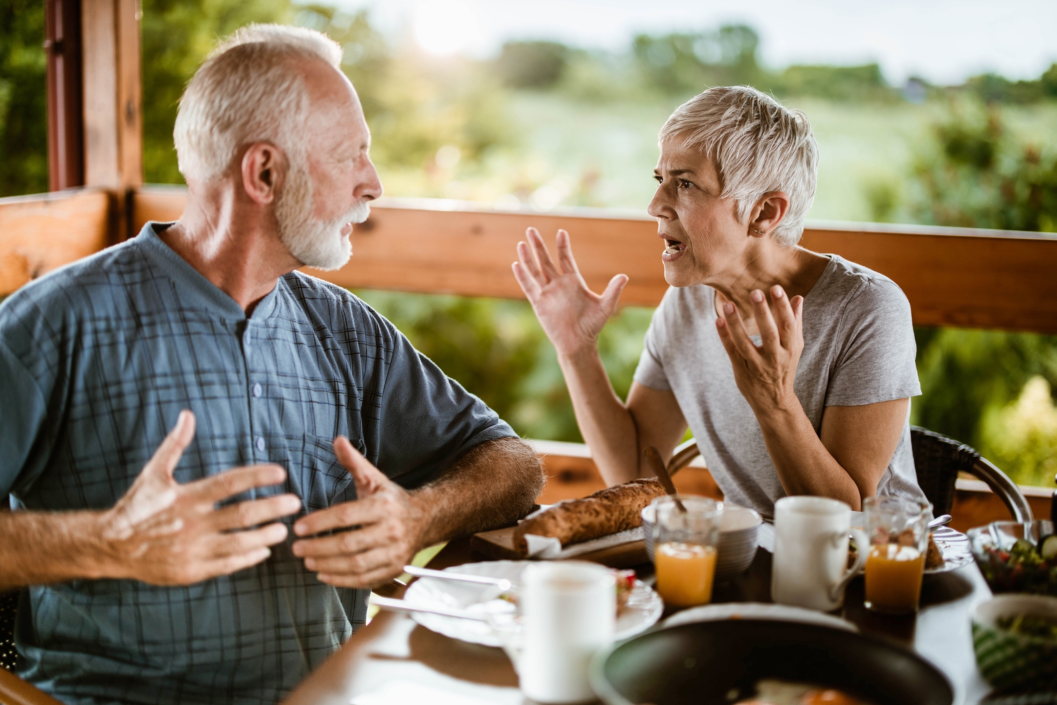Older couple having an animated conversation at a table with breakfast items, including juice and coffee, on a porch with greenery in the background