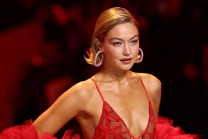 Gigi Hadid in a red gown with a deep neckline and large hoop earrings, walking on the runway at a fashion show