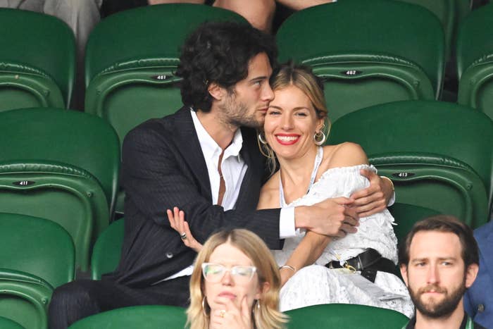 Two people sit closely in a stadium, one in a dark suit and the other in a white off-the-shoulder dress, looking happy and relaxed