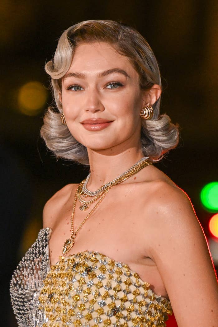Gigi Hadid in glamorous, sequined strapless gown with sleek hairstyle and layered gold necklaces, posing on a red carpet