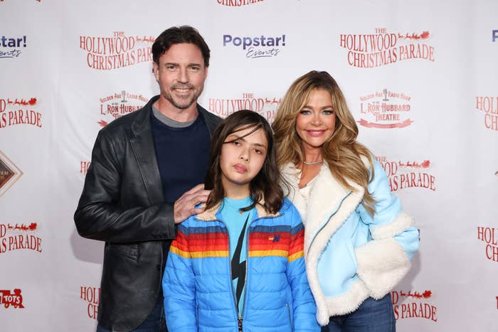 Three people at a Hollywood event, one in a jacket and two in stylish outerwear, pose on a red carpet with a backdrop of event logos