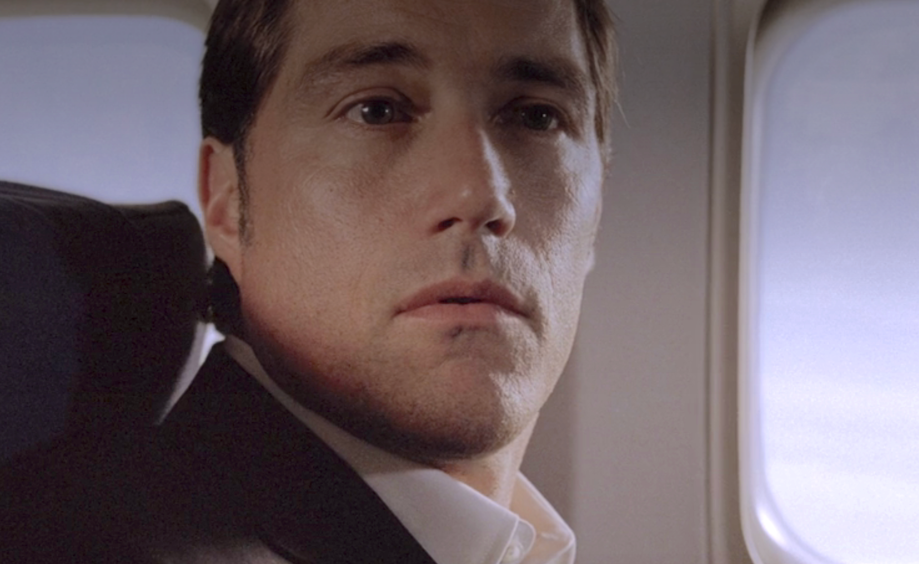 Jack on a plane, looking thoughtful, seated by a window. Visible background shows a sky view