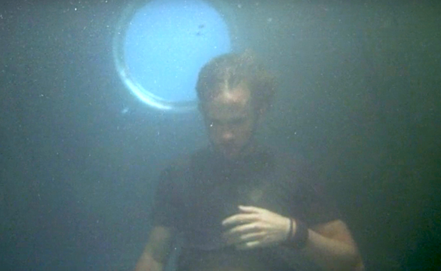 Charlie underwater near a circular window, looking down with a hand on their chest