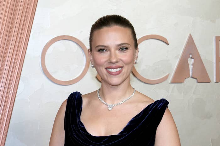 A person is on a red carpet, smiling, wearing an elegant, sleeveless dress with a necklace