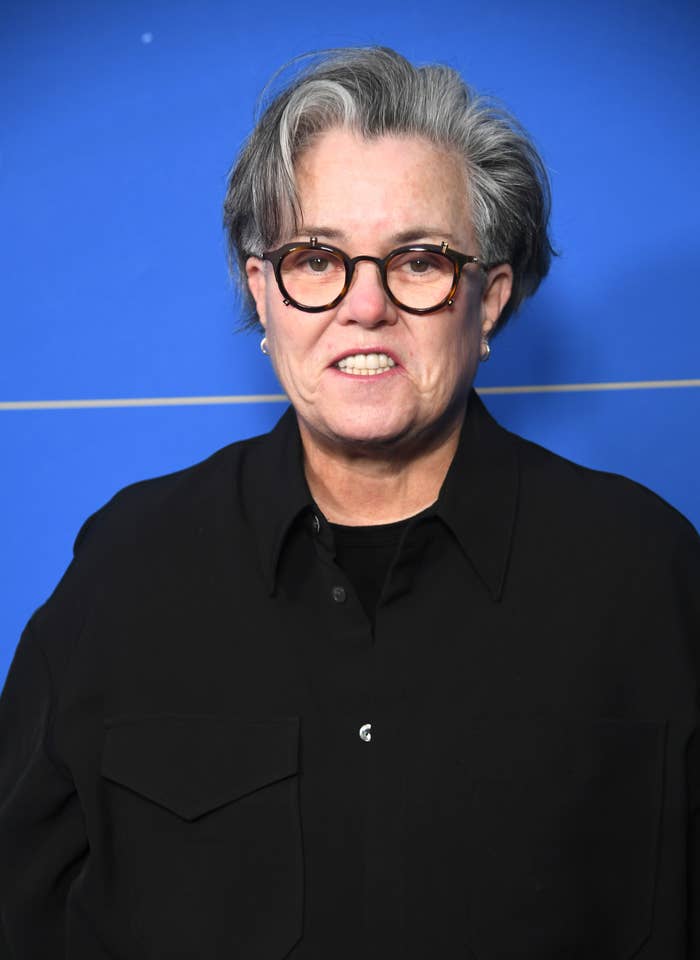 Rosie O'Donnell with short hair and glasses, wearing a black shirt, posing against a plain backdrop
