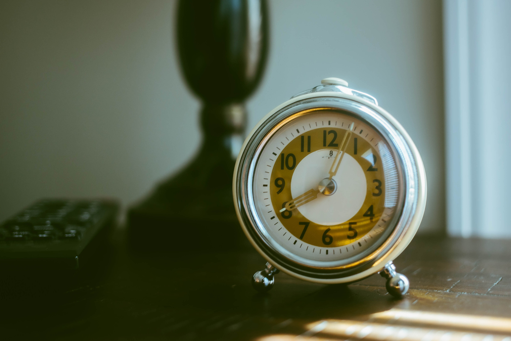 A vintage round alarm clock sits on a wooden table with a lamp and a remote nearby
