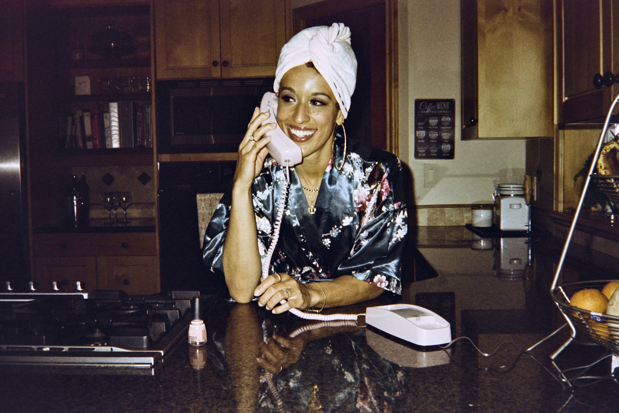Person in a floral robe with a towel on head smiles while talking on a corded phone in a kitchen