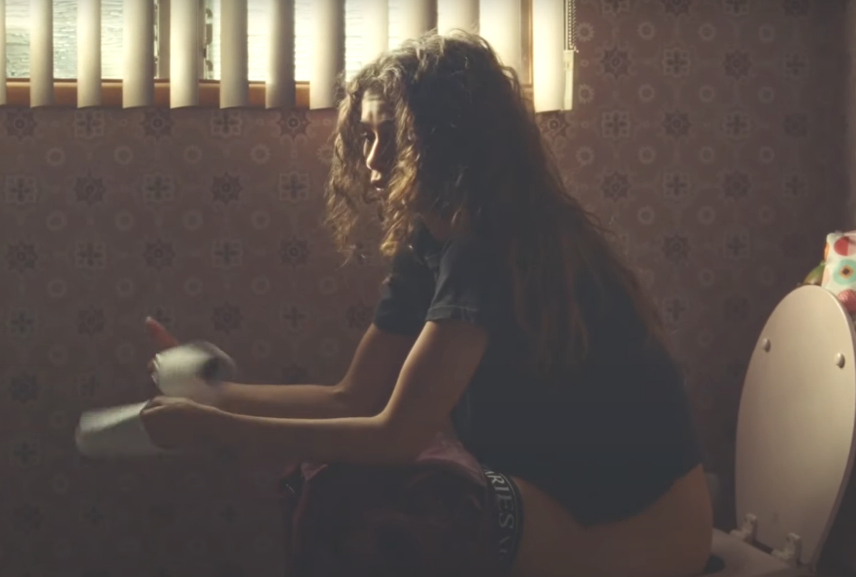 Person sitting on a toilet in a dimly lit room, holding toilet paper, with blinds partially open above