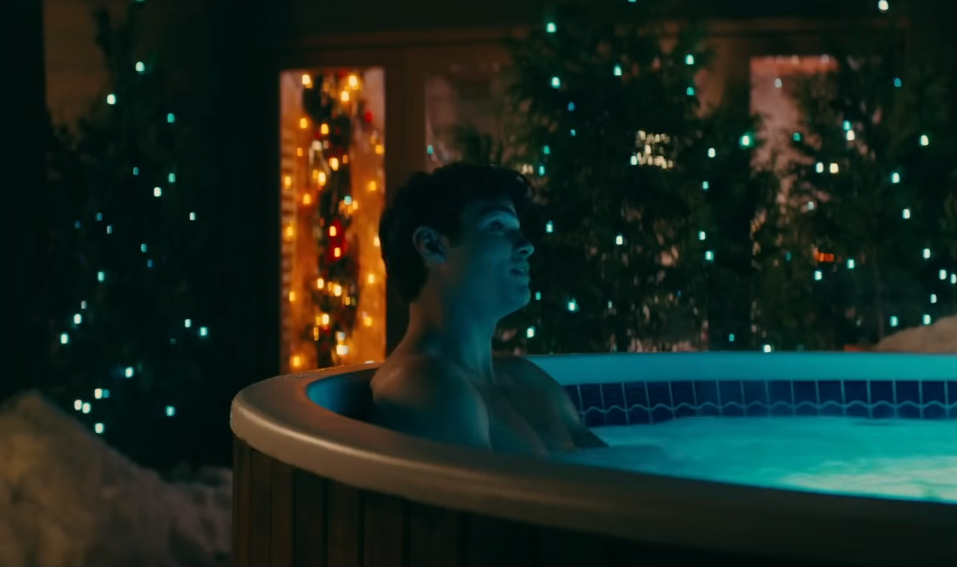 A person relaxes in a hot tub at night, surrounded by trees and festive lights. The setting suggests a cozy, holiday atmosphere