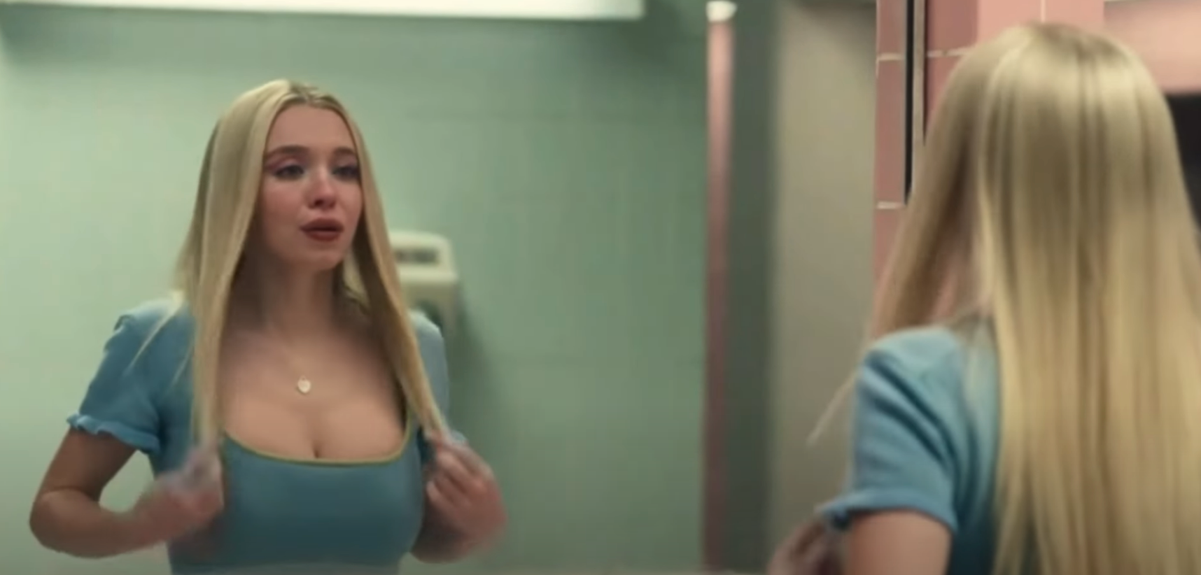 Person with long hair looks in a bathroom mirror, appearing emotional, wearing a fitted top