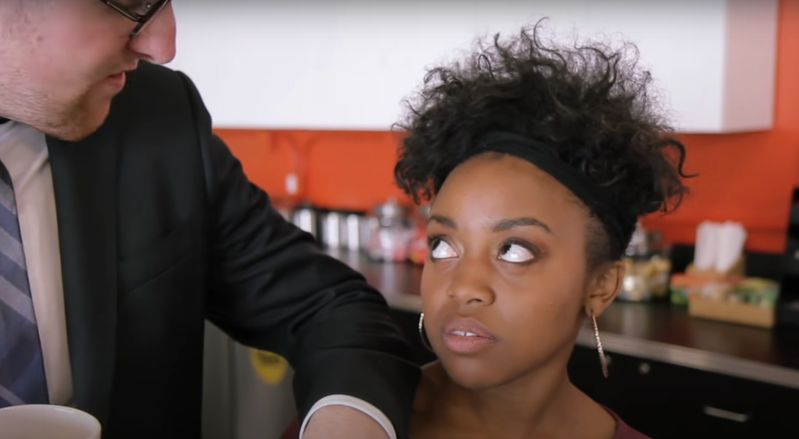 A man in a suit looks down at a woman with an annoyed expression in a kitchen setting