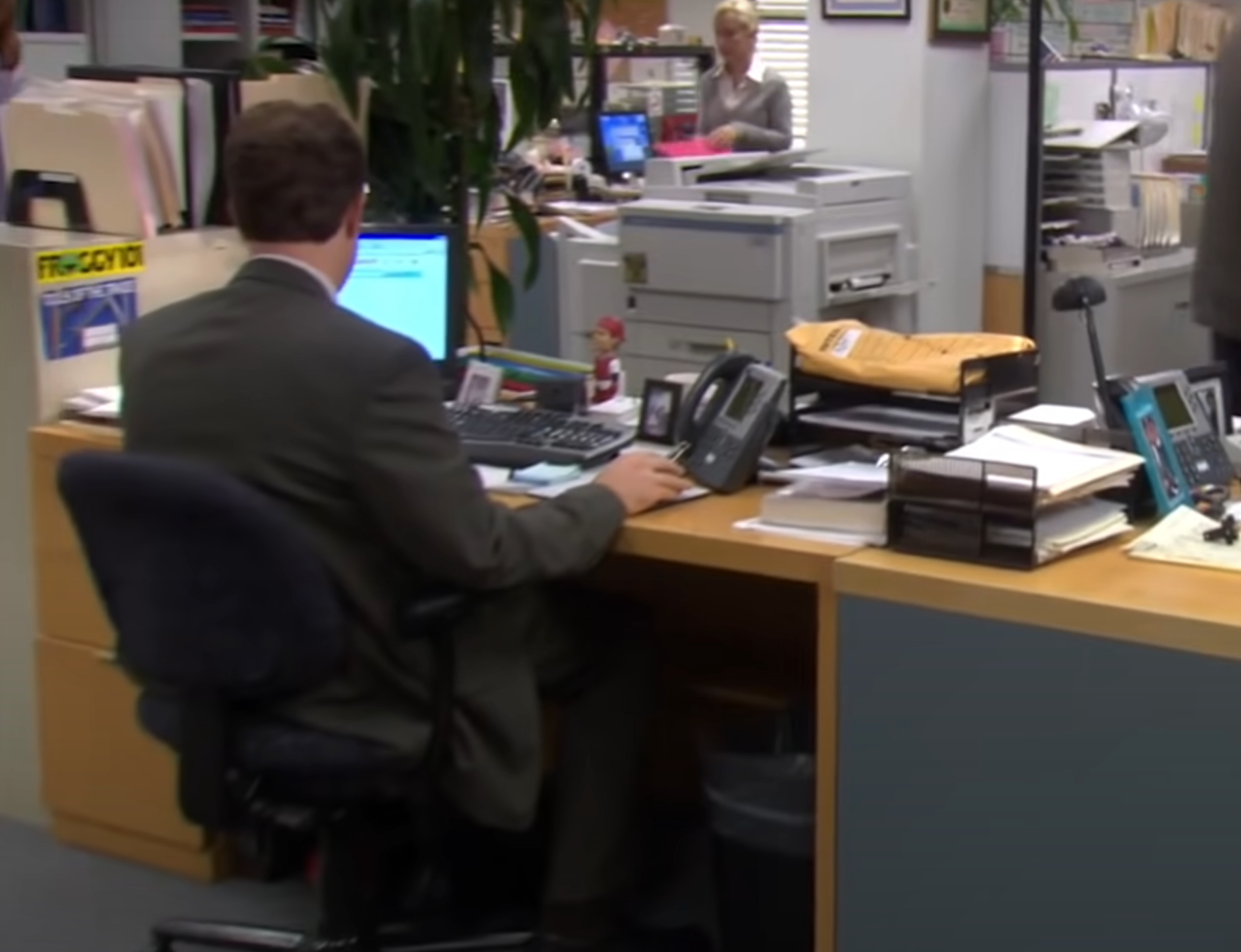 Person at an office desk using a computer; the workspace is cluttered with papers. Background shows an office setting with a coworker