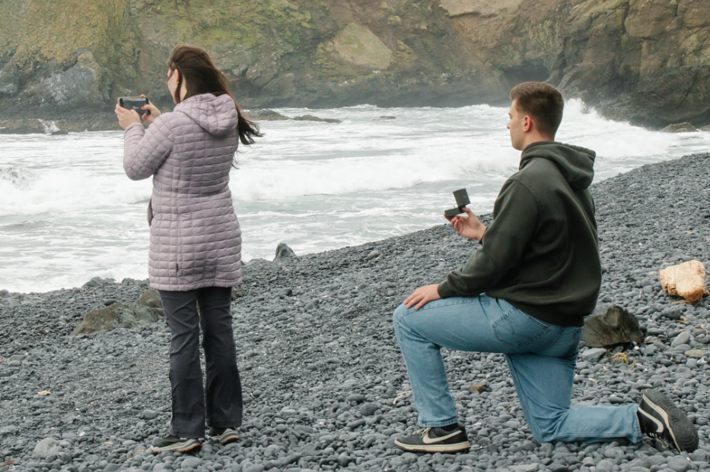 女性がカメラで海鳥を撮影し、男性がひざまずいているユーモラスな瞬間。テキストあり。