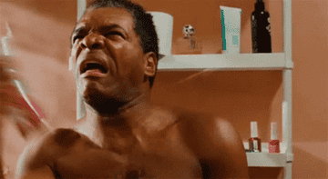 A man in a bathroom reacts playfully to using a spray can, with shelves of toiletries visible behind him