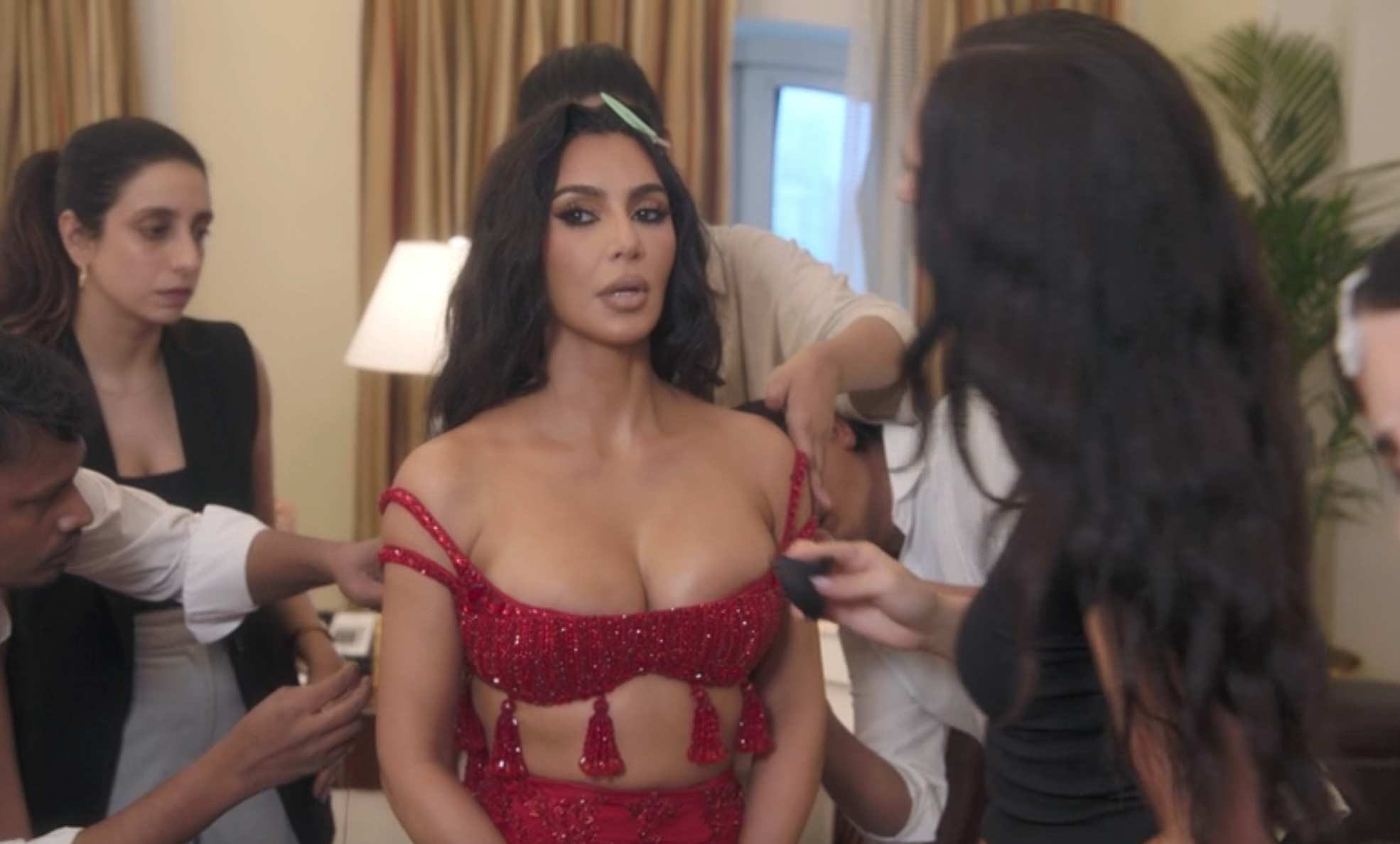 Kim Kardashian in an ornate, red beaded outfit being attended to by a team, preparing for an event