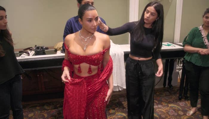 Kim wearing a red embellished outfit being adjusted by stylists in a dressing room