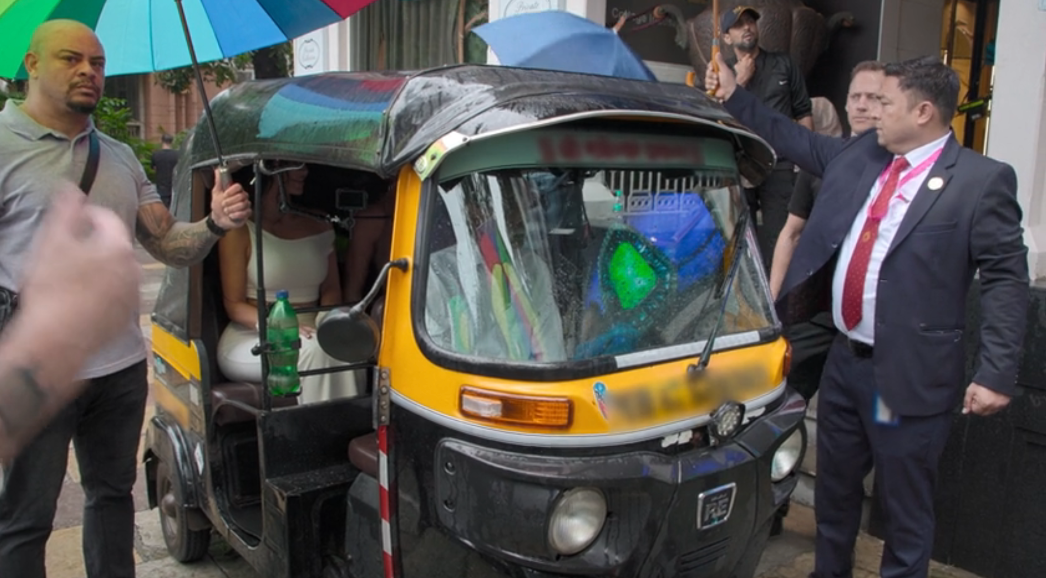Kim and Khloe sitting inside an auto rickshaw on a city street, surrounded by people, including one holding an umbrella