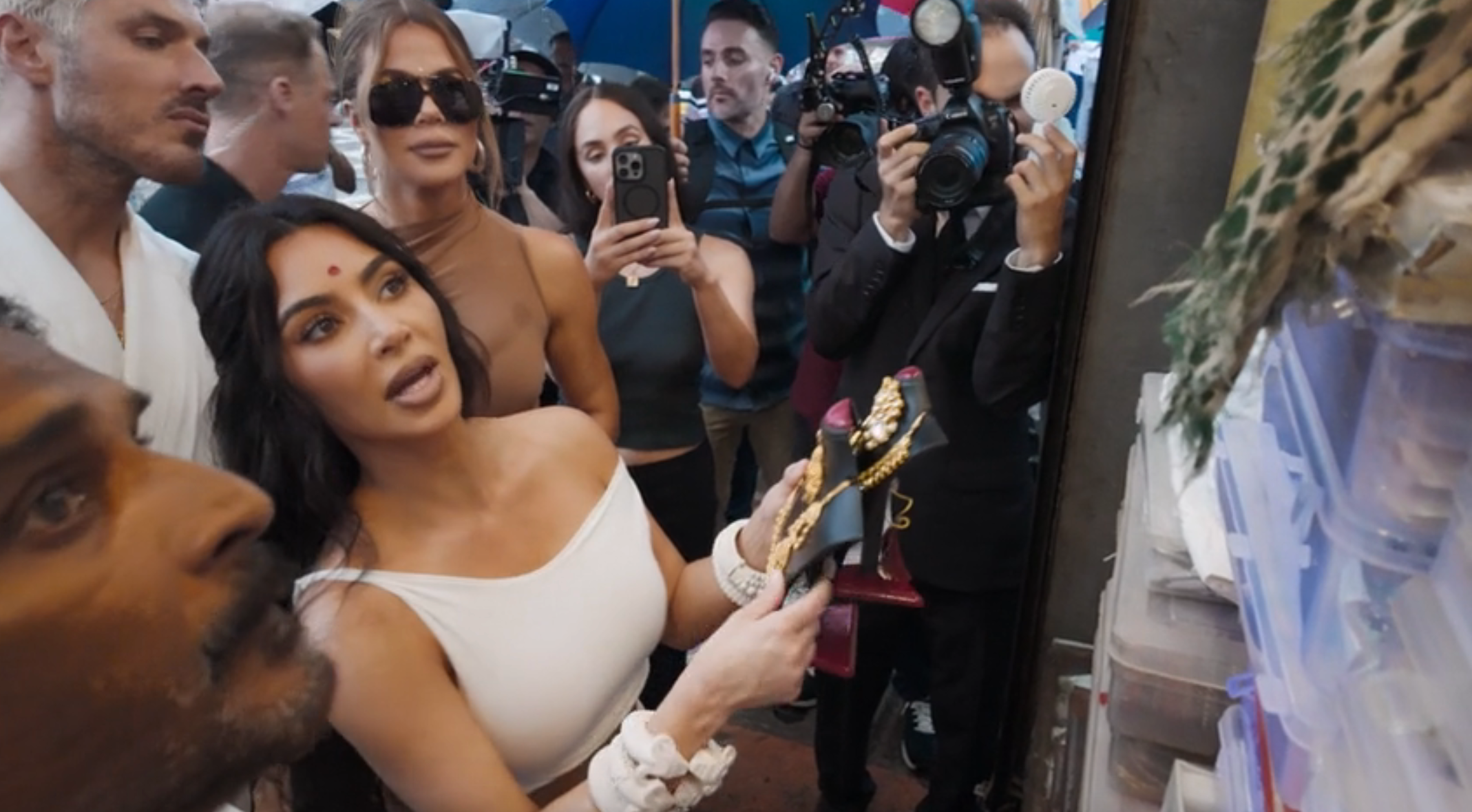 Kim Kardashian wearing a stylish white dress looks at a market stall as a crowd of people take photos with their phones