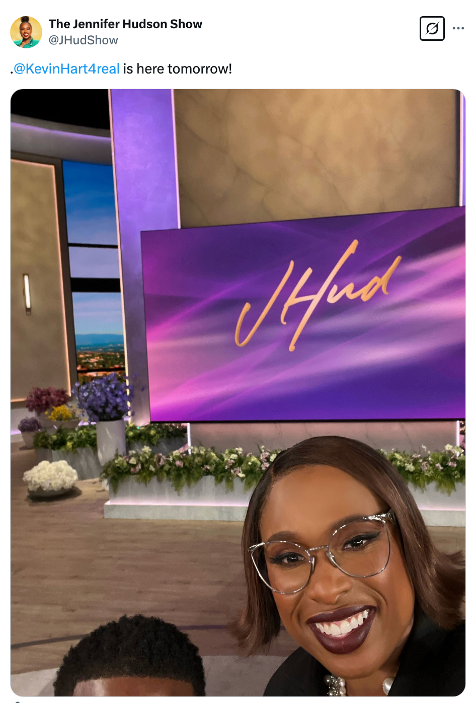 A person taking a selfie on a TV set with a screen displaying "JHud." Another person partially visible. Text mentions a guest appearing tomorrow