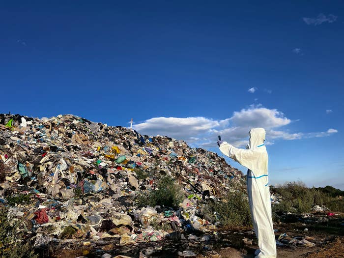 Person in protective suit stands near a large pile of waste, taking a photo. Clear blue sky in the background