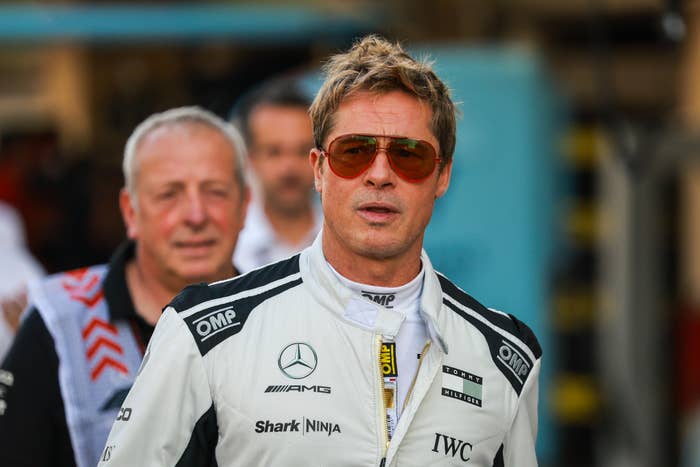 A man in a racing jumpsuit with logos walks confidently on a racetrack, wearing aviator sunglasses; a person in the background