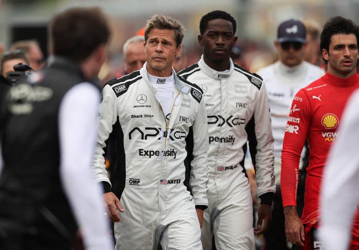 Actors in racing suits walk on a racetrack, accompanied by race car drivers. The suits feature various sponsor logos