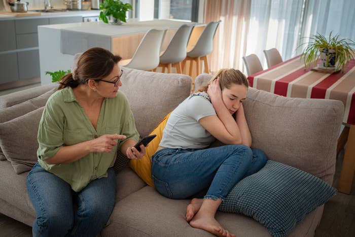 A woman on a couch holds a phone and talks to a young girl curled up next to her, looking upset. They are in a living room setting