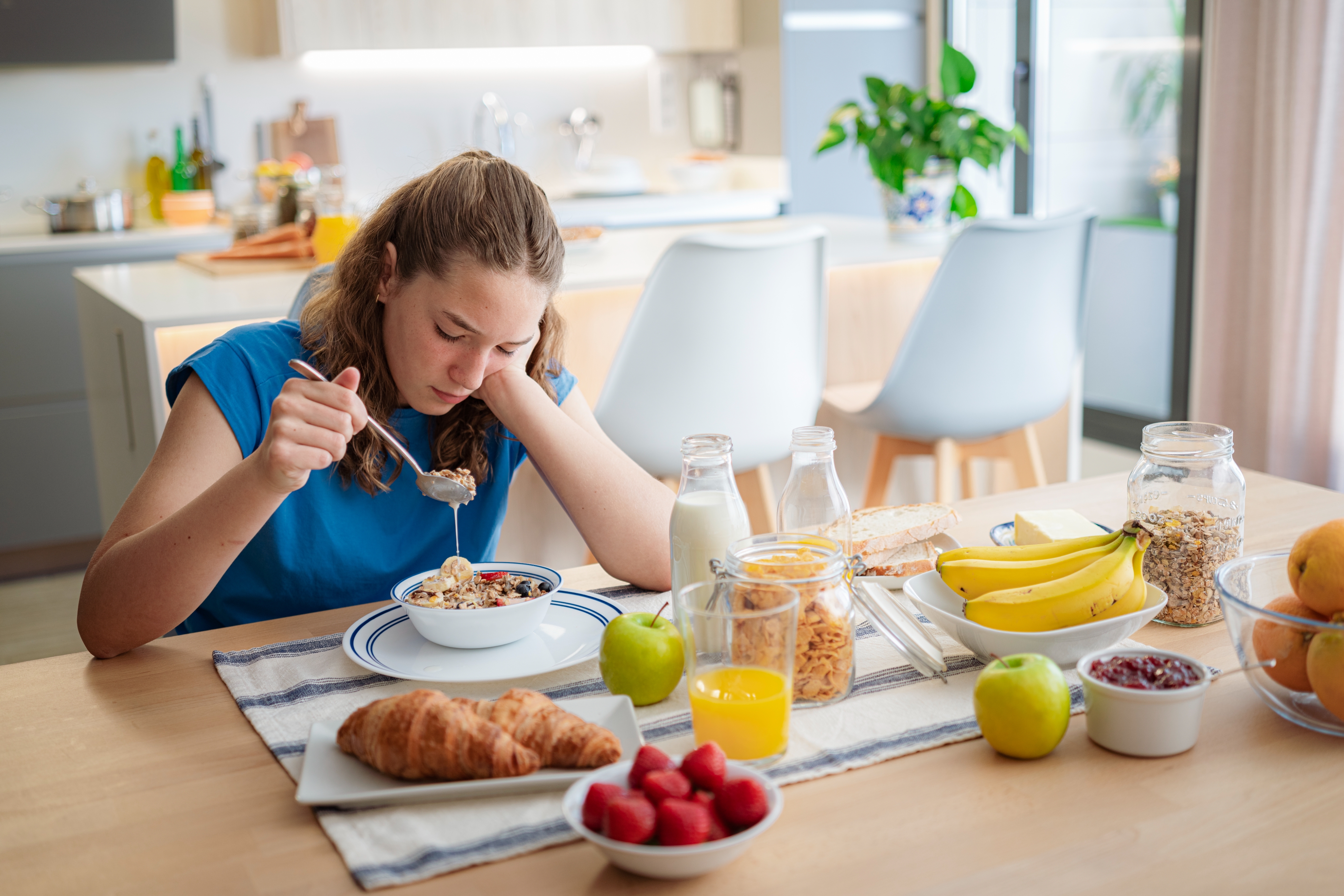 Person eating breakfast at a kitchen table, with cereal, fruit, and drinks laid out
