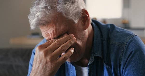 A person with gray hair sits indoors, covering their face with one hand, appearing upset or deep in thought