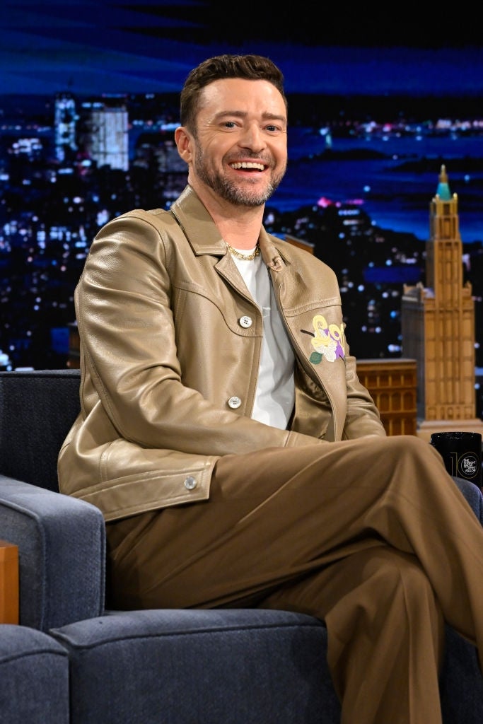 Person on a talk show set wearing a stylish leather jacket and trousers, sitting with legs crossed, smiling. Cityscape backdrop visible behind