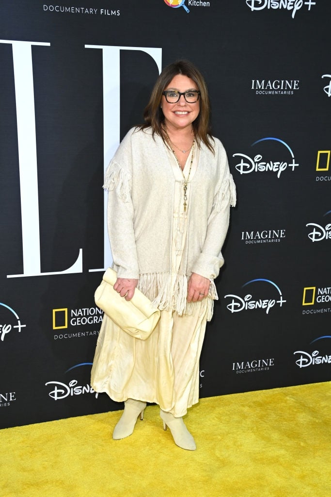 Person in layered, casual cream outfit on red carpet, holding a clutch. Various logos in the background indicate entertainment and documentary brands