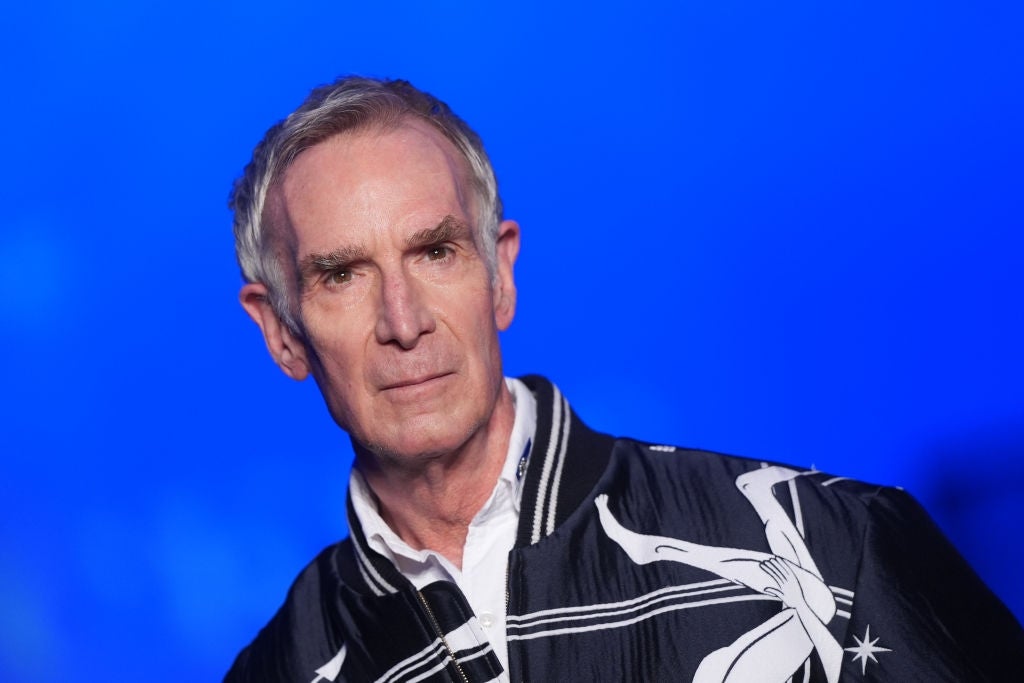 Person in a white shirt and patterned jacket stands against a blue background with a neutral expression