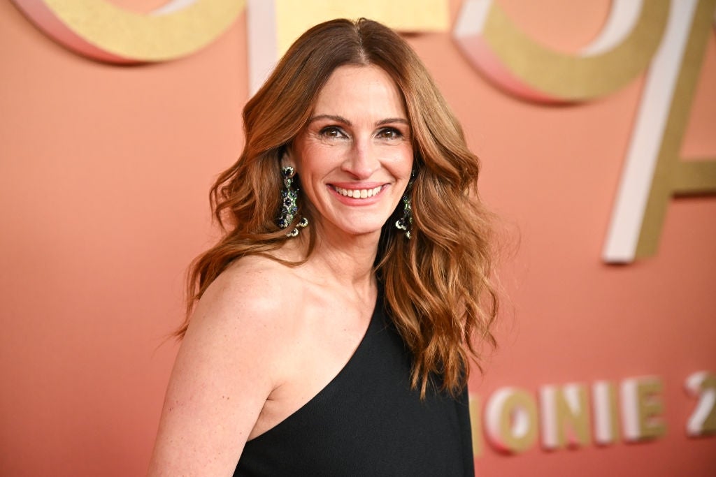 Person in elegant, one-shoulder dress smiles on a red carpet, wearing statement earrings