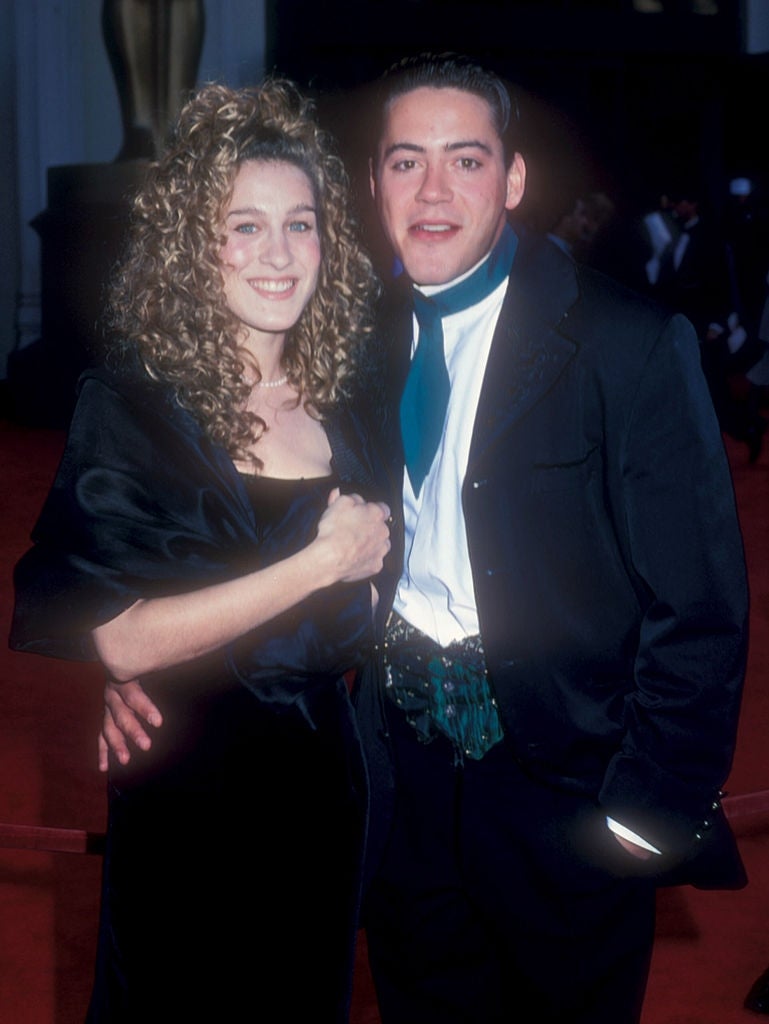 Two individuals on a red carpet; one in a velvet dress with curly hair, the other in a suit with a patterned vest