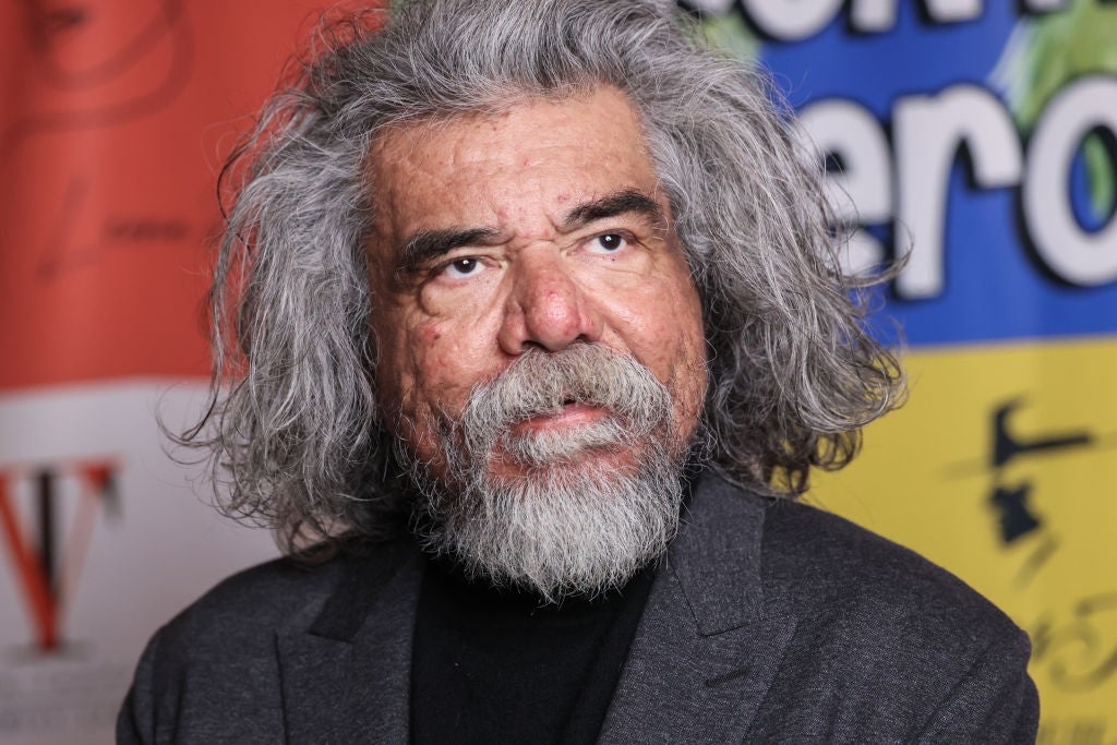 Elderly man with long, gray hair and beard in a suit, standing in front of colorful banners