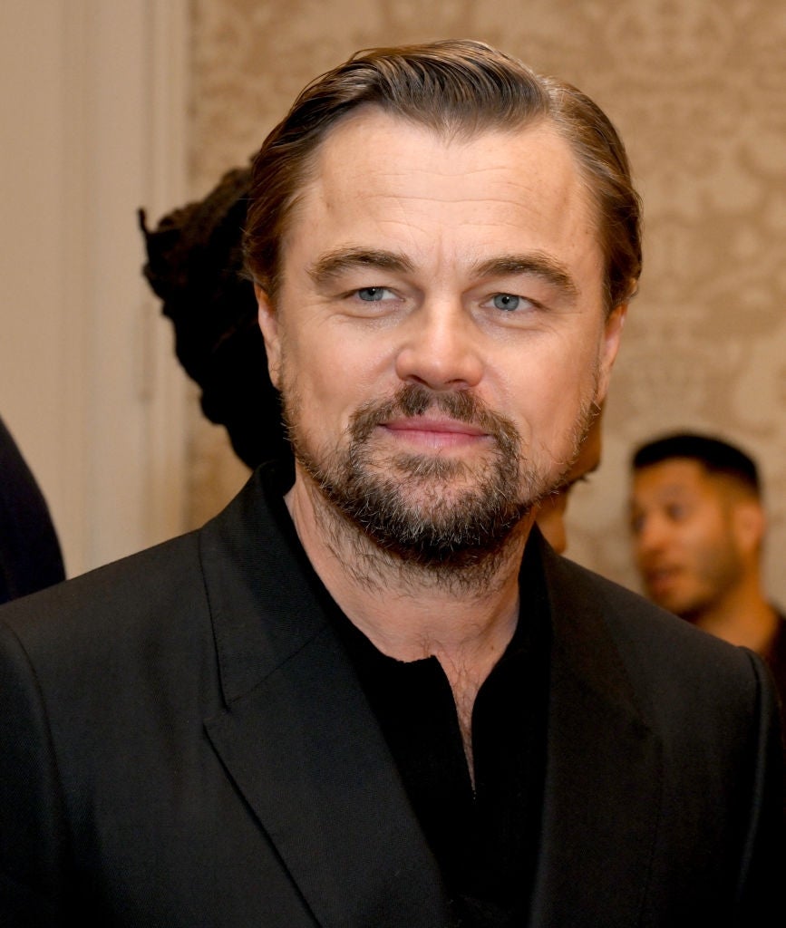 Person in formal attire at an indoor event, wearing a black suit with a subtle smile against a patterned background