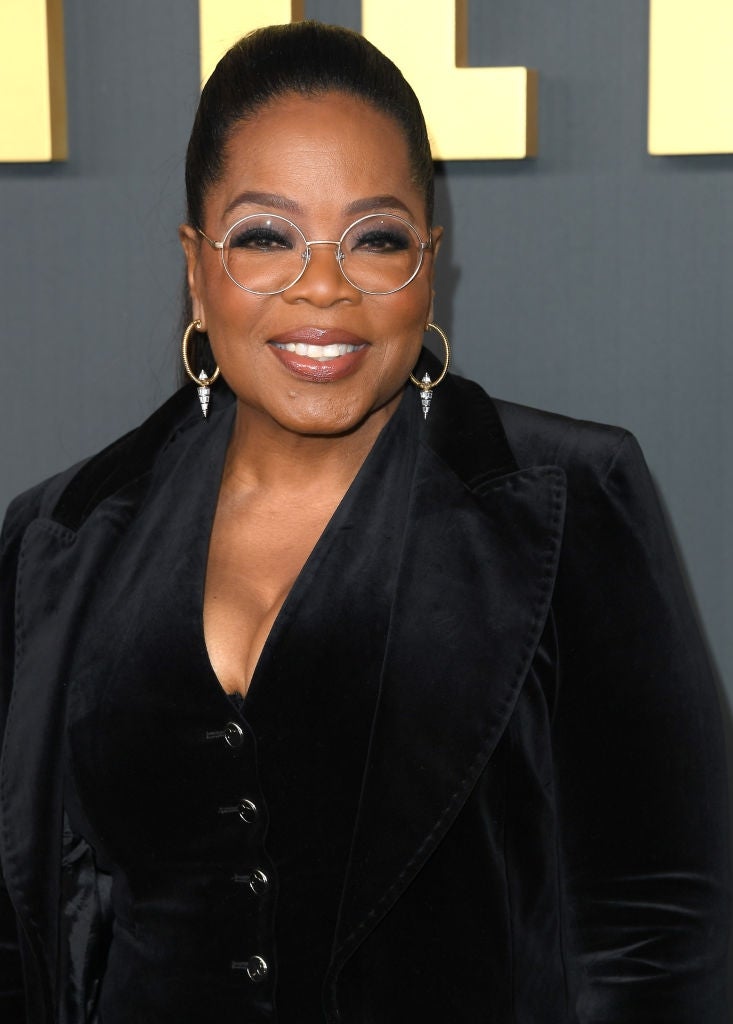 Person smiling at event, wearing velvet jacket with buttoned top, hoop earrings, and round glasses. Hair is styled back