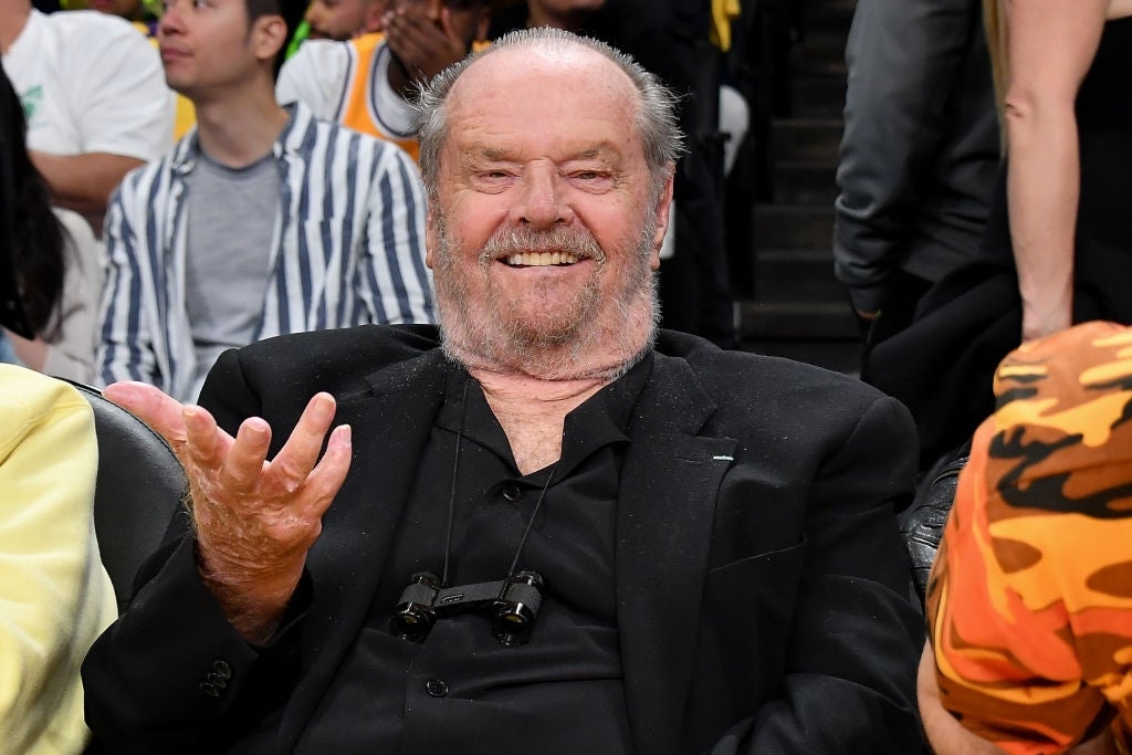 Man in a suit smiling while seated at an event, gesturing with one hand. People and seats visible in the background