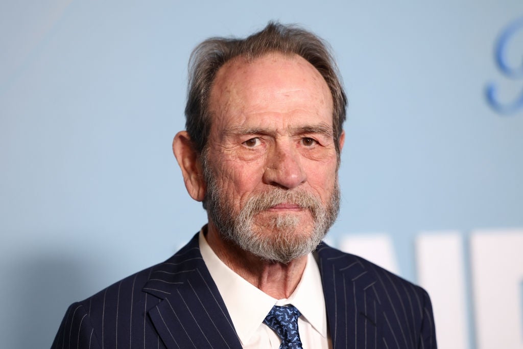 An older man with a beard in a pinstripe suit and patterned tie stands against a plain background at an event