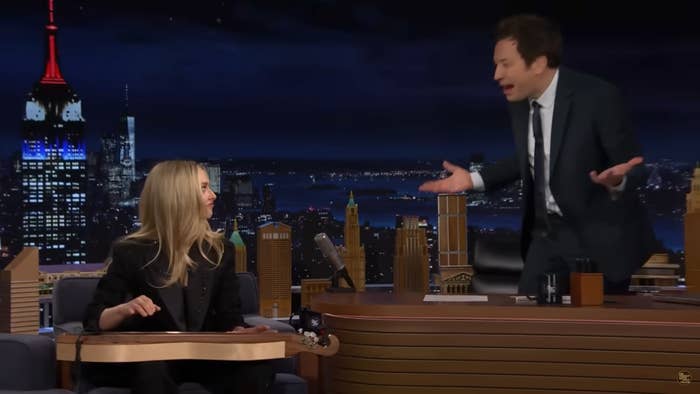 Jimmy Fallon in a smart suit animatedly talks to Amanda Seyfried on a TV talk show set resembling a nighttime city skyline