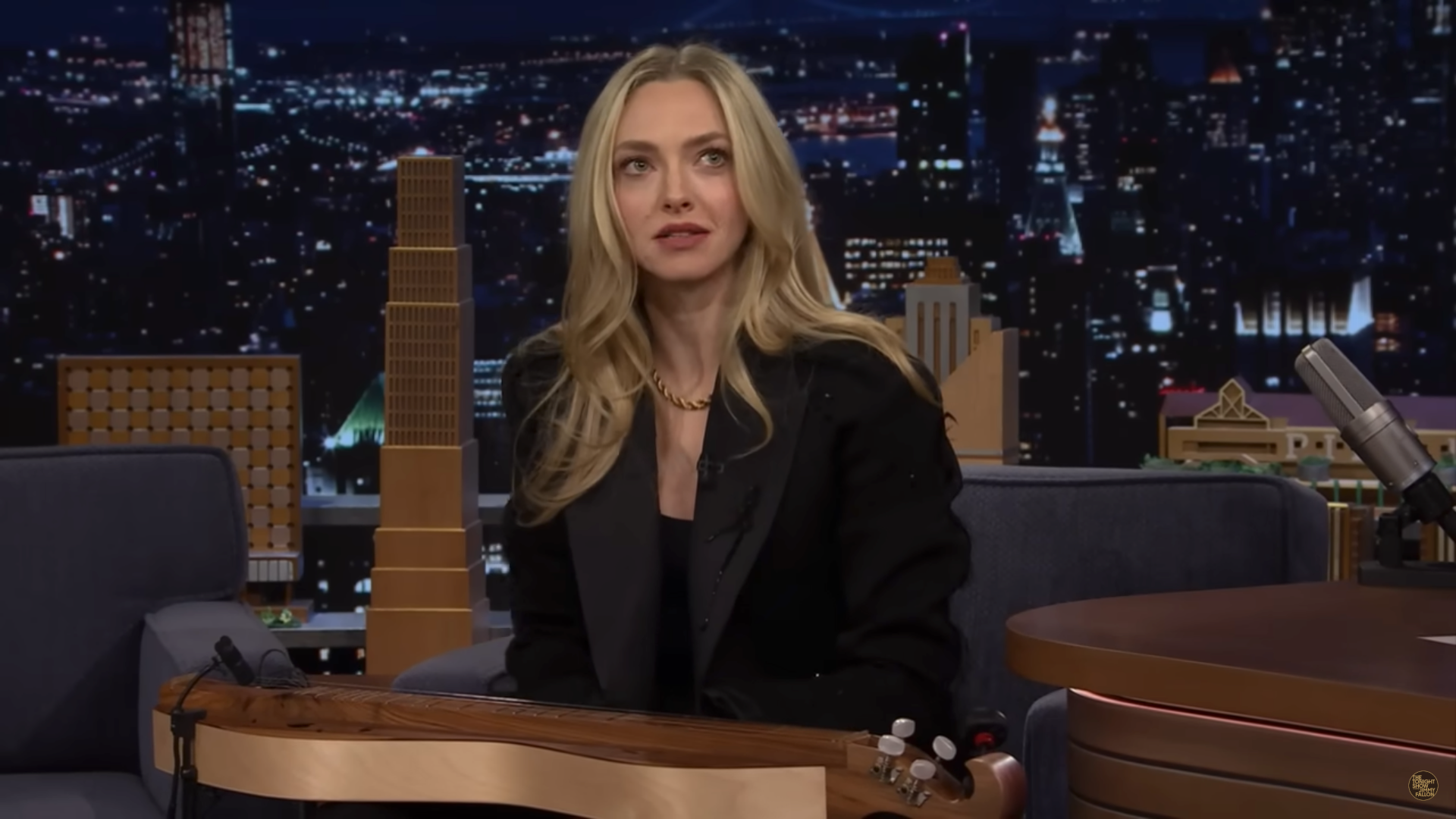 Amanda Seyfried in a sleek black jacket sits on a talk show set with a city skyline backdrop