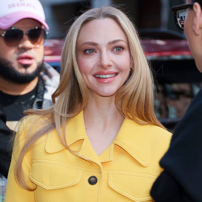 Amanda Seyfried with long hair smiling, wearing a stylish buttoned jacket, speaking to someone