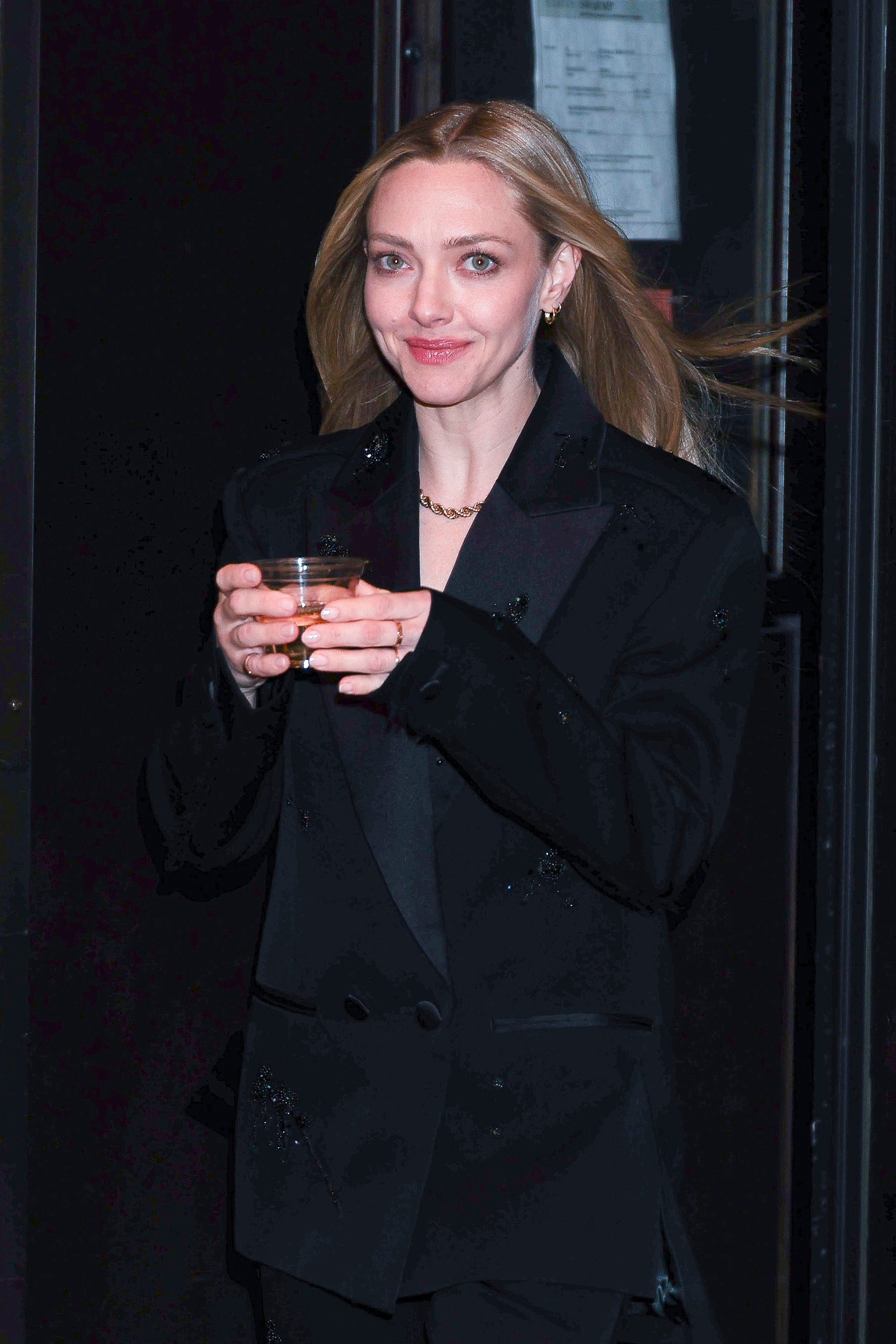 Person in a sleek black blazer holds a drink, smiling while looking at the camera