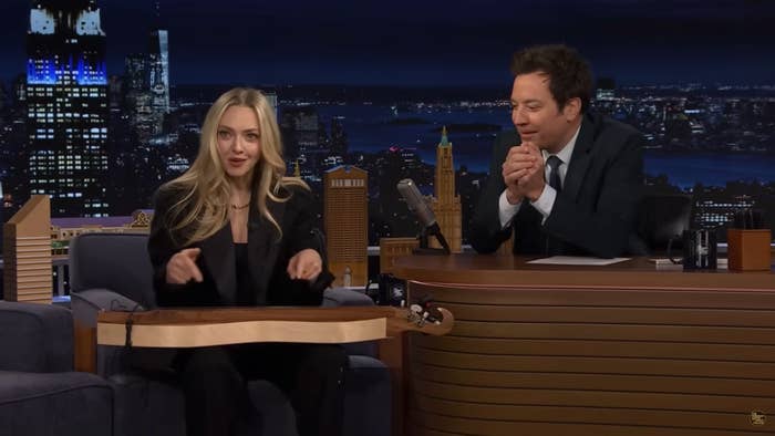 Amanda Seyfried host on a nighttime talk show, seated at a desk with city skyline in the background