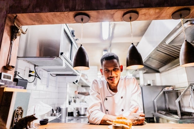 Chef in professional kitchen, wearing a double-breasted chef coat, leans on counter with a confident expression
