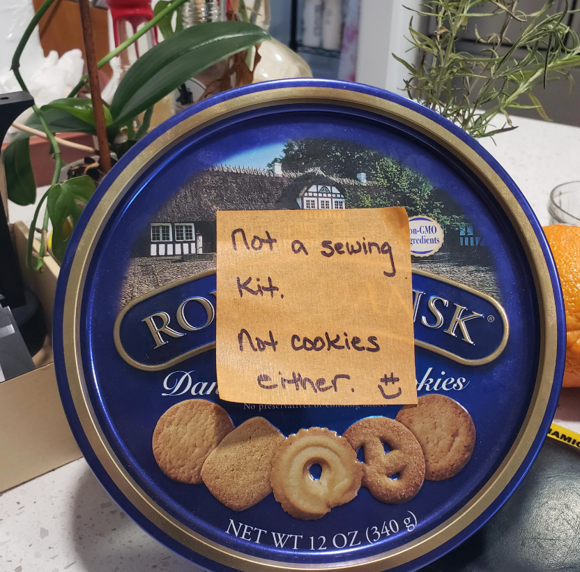 Round tin with various cookies pictured. A sticky note on it reads: "Not a sewing kit. Not cookies either. :)"